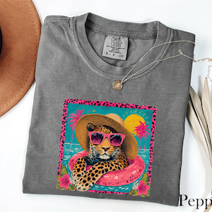 May include: A gray Comfort Colors t-shirt featuring a leopard wearing sunglasses and a straw hat, floating in a pink inner tube. The graphic is framed with a pink leopard print border and tropical imagery. A necklace and a brown hat are also visible.
