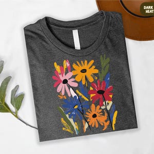 a gray t-shirt with a colorful floral design, including yellow, pink, orange, and blue flowers, along with green leaves. The t-shirt is placed on a white surface, and there is a brown hat visible in the background.