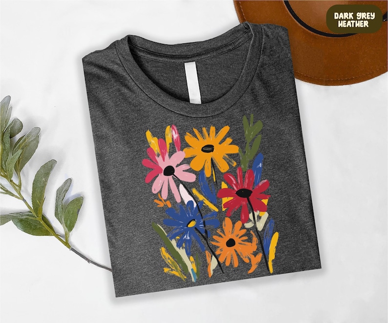 a gray t-shirt with a colorful floral design, including yellow, pink, orange, and blue flowers, along with green leaves. The t-shirt is placed on a white surface, and there is a brown hat visible in the background.
