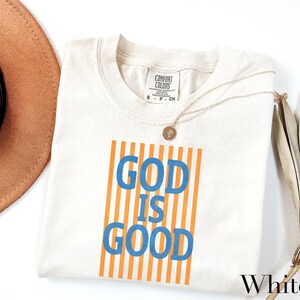 A white t-shirt with the text "GOD IS GOOD" printed on it, along with a necklace and a brown hat.