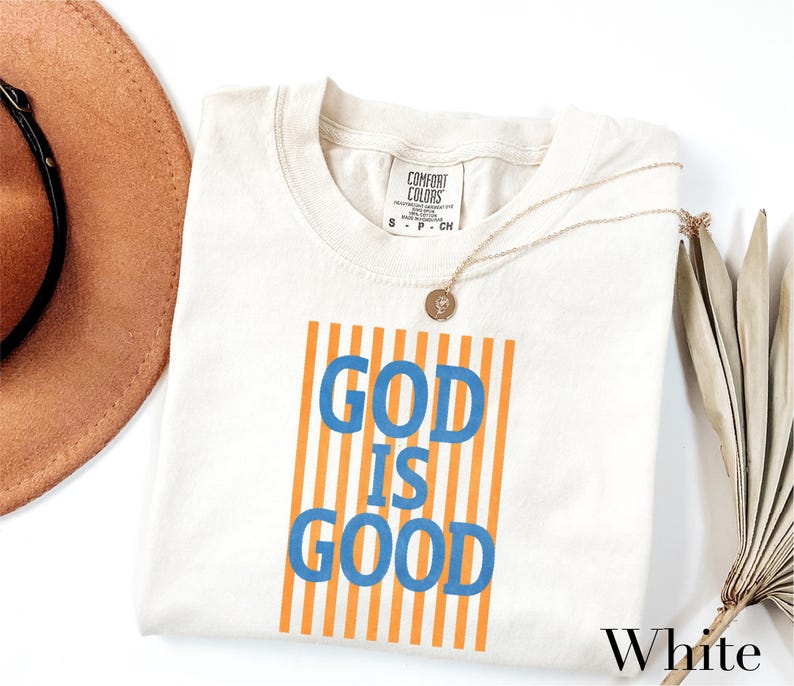 A white t-shirt with the text "GOD IS GOOD" printed on it, along with a necklace and a brown hat.