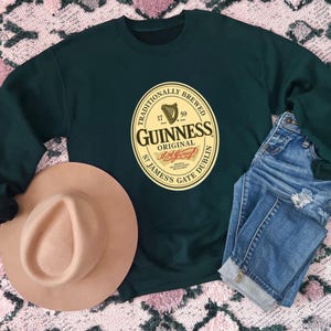 a dark green sweatshirt with a Guinness logo, a tan hat, and a pair of blue jeans laid out on a patterned surface.