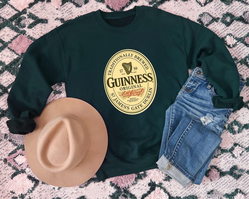 a dark green sweatshirt with a Guinness logo, a tan hat, and a pair of blue jeans laid out on a patterned surface.