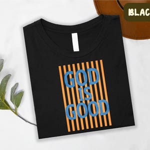 A black t-shirt with the text "GOD IS GOOD" printed on it, along with orange and blue vertical stripes.
