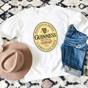 a white sweatshirt with a Guinness logo, a brown hat, and a pair of blue jeans laid out on a patterned rug.