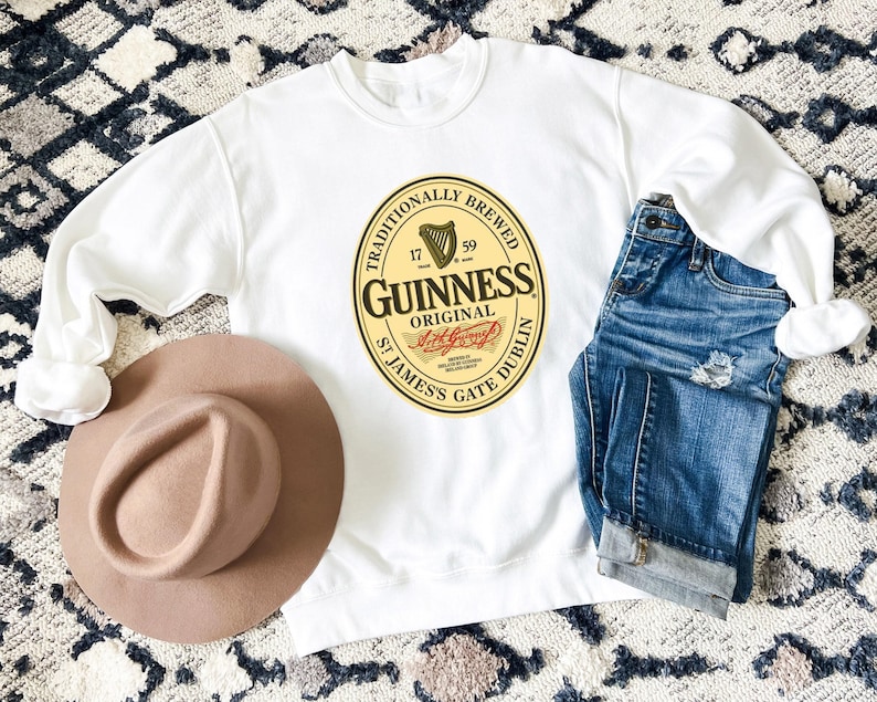 a white sweatshirt with a Guinness logo, a brown hat, and a pair of blue jeans laid out on a patterned rug.