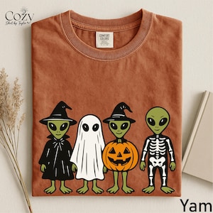 May include: A rust-colored crew neck t-shirt featuring a graphic of four green aliens in Halloween costumes: one as a witch, one as a ghost, one inside a jack-o'-lantern, and one as a skeleton. The shirt has a Comfort Colors brand neck label.