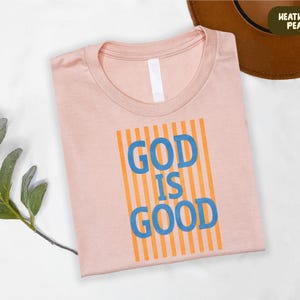 A pink t-shirt with the text "GOD IS GOOD" printed on it, along with a brown hat and some greenery.