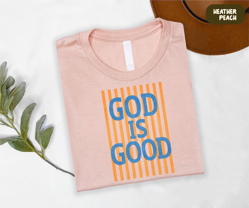 A pink t-shirt with the text "GOD IS GOOD" printed on it, along with a brown hat and some greenery.