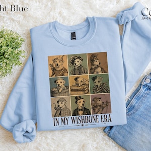 A light blue sweatshirt with a graphic design of nine different dog illustrations arranged in a 3x3 grid, along with the text "IN MY WISHBONE ERA" printed below.