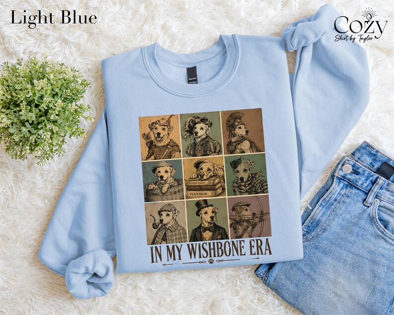 A light blue sweatshirt with a graphic design of nine different dog illustrations arranged in a 3x3 grid, along with the text "IN MY WISHBONE ERA" printed below.