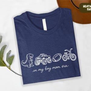 A blue t-shirt with white text and illustrations of a dinosaur, truck, bicycle, and football, along with the phrase "in my boy mom era" printed on it.