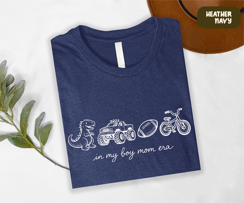 A blue t-shirt with white text and illustrations of a dinosaur, truck, bicycle, and football, along with the phrase "in my boy mom era" printed on it.