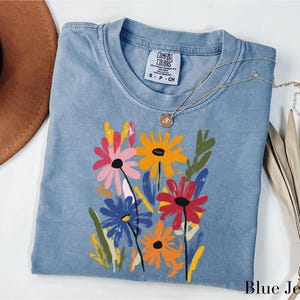 A blue denim t-shirt with a colorful floral design, a brown hat, and a necklace are arranged on a white surface.
