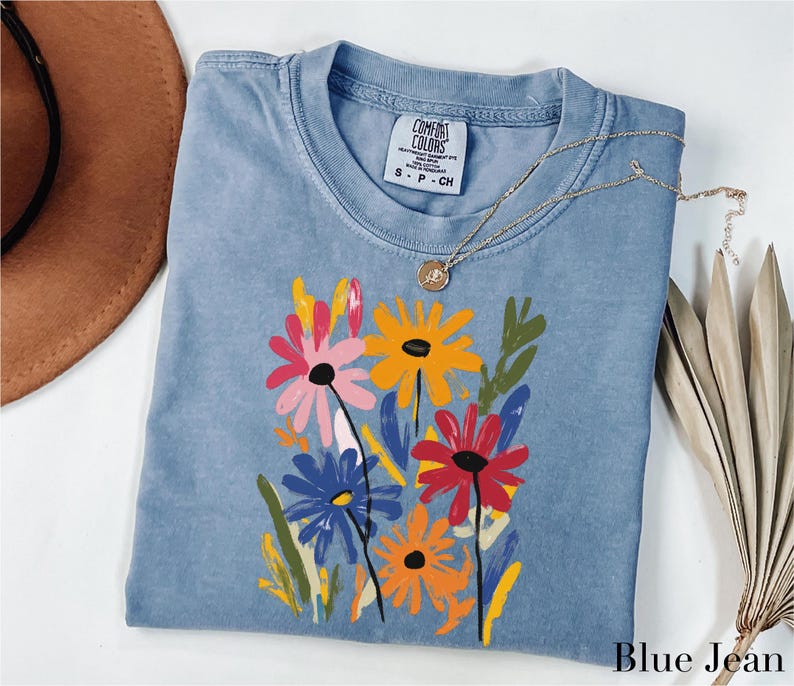 A blue denim t-shirt with a colorful floral design, a brown hat, and a necklace are arranged on a white surface.