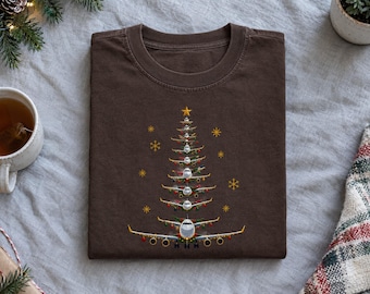 Puede incluir: Una camiseta de cuello redondo de color marrón oscuro con un gráfico de un árbol de Navidad formado por aviones apilados. El diseño incluye pequeñas luces de colores, detalles de estrellas doradas y una estrella amarilla en la cima.