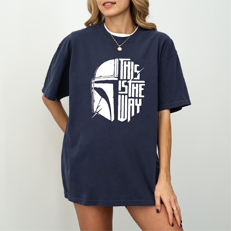 A woman wearing a navy blue t-shirt with a graphic design featuring a helmet and the text "THIS IS THE WAY" printed on it.