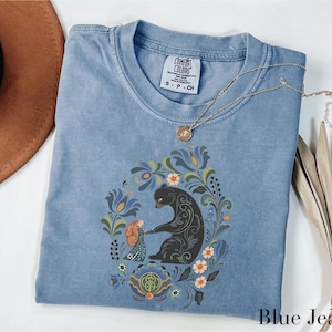 May include: A blue jean colored t-shirt with a floral and bear graphic. The graphic features a bear and a person holding hands, surrounded by flowers and leaves. The shirt has a crew neck and short sleeves. A gold necklace is draped over the shirt.