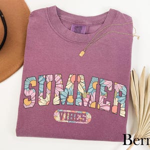 A purple t-shirt with the word "SUMMER" printed on it, along with a necklace and a hat, all placed on a white background.