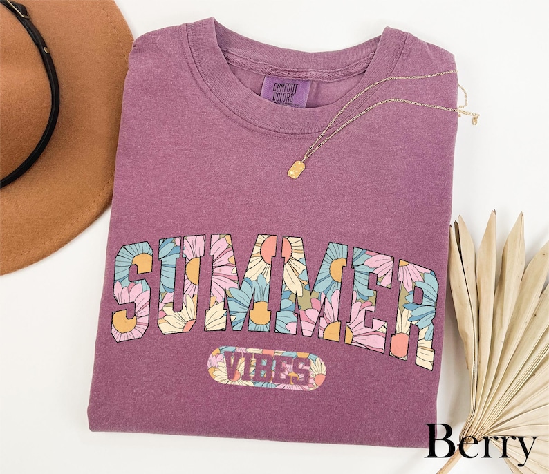 A purple t-shirt with the word "SUMMER" printed on it, along with a necklace and a hat, all placed on a white background.