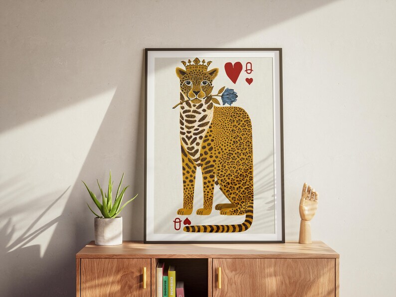 May include: A framed art print featuring a stylized leopard wearing a crown and holding a blue rose. The leopard has a spotted coat and is depicted with playing card symbols, including the Queen of Hearts. The artwork is displayed on a wooden cabinet.