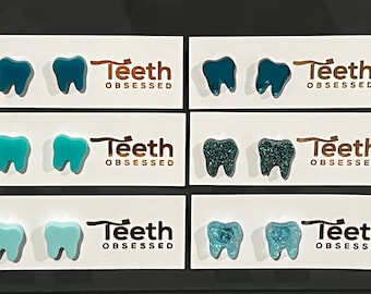 Peut inclure: Six paires de boucles d'oreilles en forme de dent sur des cartes blanches. Les boucles d'oreilles sont en résine et sont disponibles en trois couleurs : sarcelle, bleu clair et sarcelle avec des paillettes. Chaque carte porte l'inscription "Teeth Obsessed".