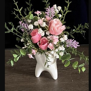 May include: A white ceramic tooth-shaped vase filled with a floral arrangement. The arrangement includes pink roses, white baby's breath, and sprigs of greenery. The vase is on a wooden surface.
