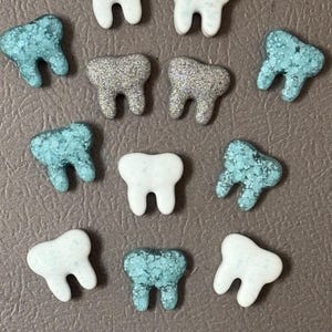 May include: An assortment of decorative tooth-shaped embellishments in various colors and textures. Some are white, others are a speckled turquoise, and two have a glittery silver finish. These small, dental-themed items are arranged on a neutral background.