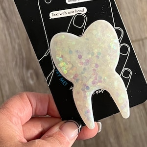 May include: White glitter phone grip shaped like a tooth. The grip says "Try Me!" and "Text with one hand" on the packaging.