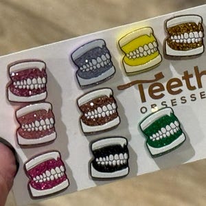 May include: A set of nine enamel pins featuring different styles of teeth, each with a glittery finish. The pins are arranged on a white card with the text "Teeth Obsessed".