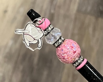 May include: Black glitter pen with pink and clear decorative beads. The pen features a charm of a cartoon figure. The pen has a black tip and a pink accent near the beads. The beads are accented with silver rhinestones.