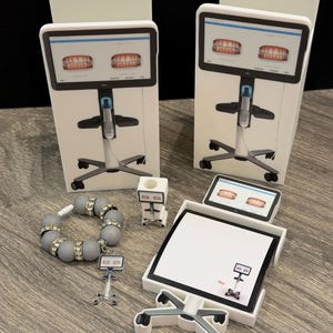 May include: A collection of miniature dental office models, including two large white displays with teeth diagrams, a bracelet with rhinestone accents, and a small chair-shaped notepad holder. The items are arranged on a wooden surface.