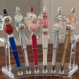 May include: A collection of decorative pens with various designs, including dental-themed charms and the text "FLOSS BOSS." The pens have colorful barrels and are displayed in a clear acrylic holder.