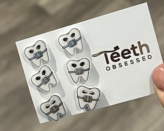 Peut inclure: Une carte blanche avec six autocollants de dents de dessin animé, chacune avec des broches. Les autocollants sont de différentes couleurs. La carte porte le texte "Teeth OBSESSED" en marron.