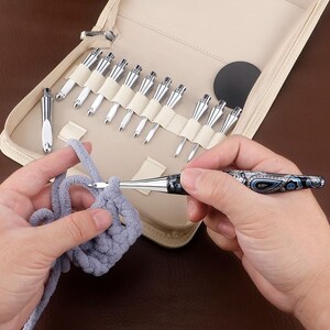May include: A set of interchangeable crochet hooks with silver metal tips and a patterned handle featuring blue, black, and white paisley designs. The hooks are stored in a beige zippered case with elastic loops, and one hook is in use with gray yarn.