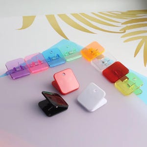 May include: A collection of square-shaped plastic clips in various colors including purple, pink, blue, green, orange, clear, red, yellow, black, and white. Each clip features a metal spring mechanism and a small circular hole at the top edge.
