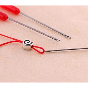 May include: Two metal beading needles with bright red plastic handles and a silver-toned metal bead featuring a black spiral design. A thin red thread is looped through the bead and attached to the tip of one of the needles for threading.