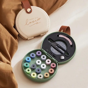 May include: A round, sage green sewing kit case with a brown leather strap and gold snap closure. The open case reveals sixteen colorful thread bobbins, black thread snips, a silver thimble, and a measuring tape. A closed beige case reads "THINGS WITH love".