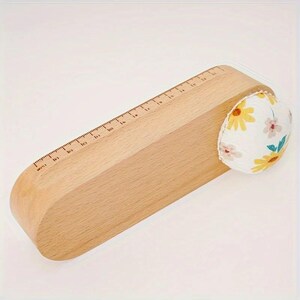 May include: A light-colored wooden sewing tool with rounded ends featuring a ruler along the top edge marked in centimeters up to 15 cm. A round, fabric-covered pincushion with a yellow and pink floral pattern is attached to one end.