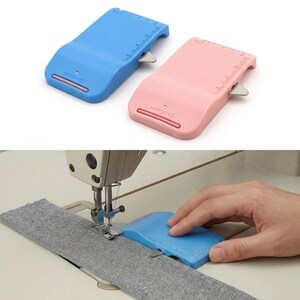 May include: A blue and a pink plastic sewing seam guide tool, each featuring a built-in metal blade and integrated ruler markings. The tool is shown being used to guide a strip of gray fabric under a sewing machine needle and presser foot.