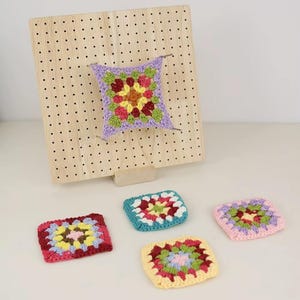May include: A wooden pegboard with a crocheted square pinned to it, and four additional colorful crocheted squares. The squares feature a floral design with various colors including red, yellow, green, and purple. The pegboard is light brown.