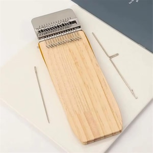 May include: A wooden darning loom tool featuring a rectangular light-colored wood base with a silver metal top component containing multiple vertical hooks. The set includes a long silver sewing needle and a thin silver metal hook tool for repair.