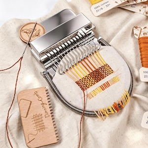 May include: A metal darning loom tool with a spring-loaded frame, featuring a woven pattern in yellow and orange thread. Includes a wooden darning comb with engraved text and a needle with brown thread, all set against a light fabric background.