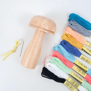 May include: A wooden mushroom-shaped thread holder with a needle and yellow threader. Colorful embroidery floss in various shades of blue, pink, green, yellow, and black are arranged on a white surface. Craft supplies for embroidery.