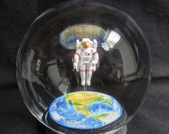 May include: A glass globe snow globe with a white astronaut figurine standing on a blue and green globe inside. The astronaut is wearing a red and white spacesuit.