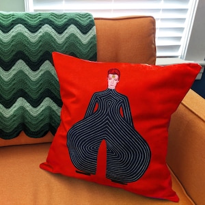 May include: Red cushion with a black and white illustration of a person wearing a patterned jumpsuit. The person has a star on their forehead.
