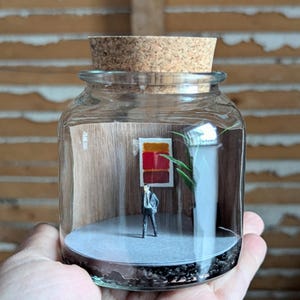 May include: A miniature scene inside a glass jar with a cork lid. The scene depicts a small figure in a suit standing in front of a red, orange and yellow abstract painting. A small potted plant is in the background.