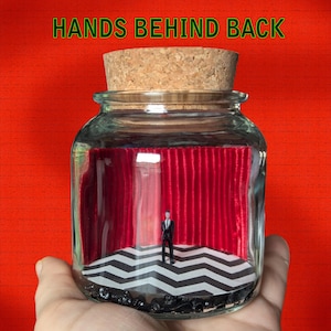 May include: A glass jar with a cork lid containing a miniature diorama of a man standing in front of a red velvet curtain and a black and white checkered floor. The text "HANDS BEHIND BACK" is printed on the top of the jar.