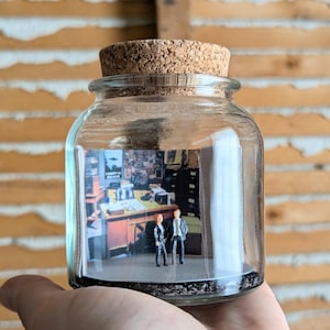 May include: A clear glass jar with a cork stopper containing a miniature diorama of an office scene. Inside, two small figurines stand on a dark base in front of a printed backdrop depicting a desk, office furniture, and a poster on the wall.