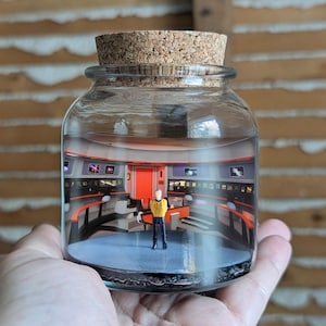 May include: A glass jar with a cork stopper, housing a miniature Star Trek diorama. Inside, a figure in a yellow uniform stands on a blue platform, surrounded by control panels and a red door. The jar contains water and black gravel.